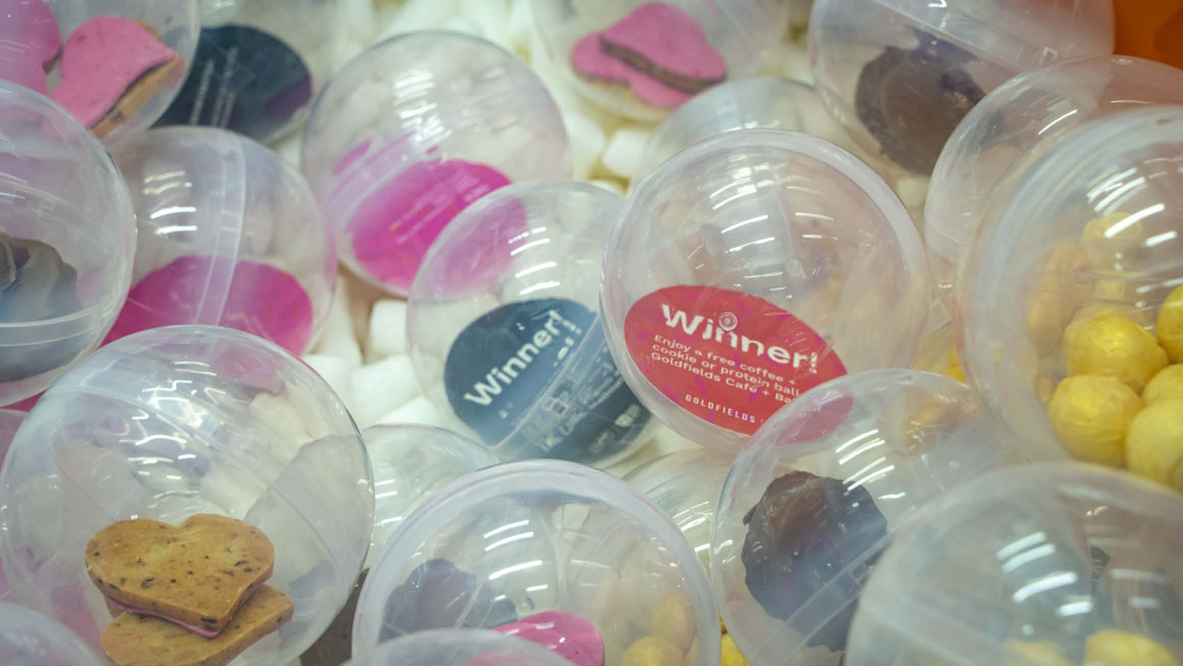 Round clear balls from a claw grab machine holding a variety of prizes, including red and black vouchers, pink heart cookies, chocolate treats, and golden round treats.