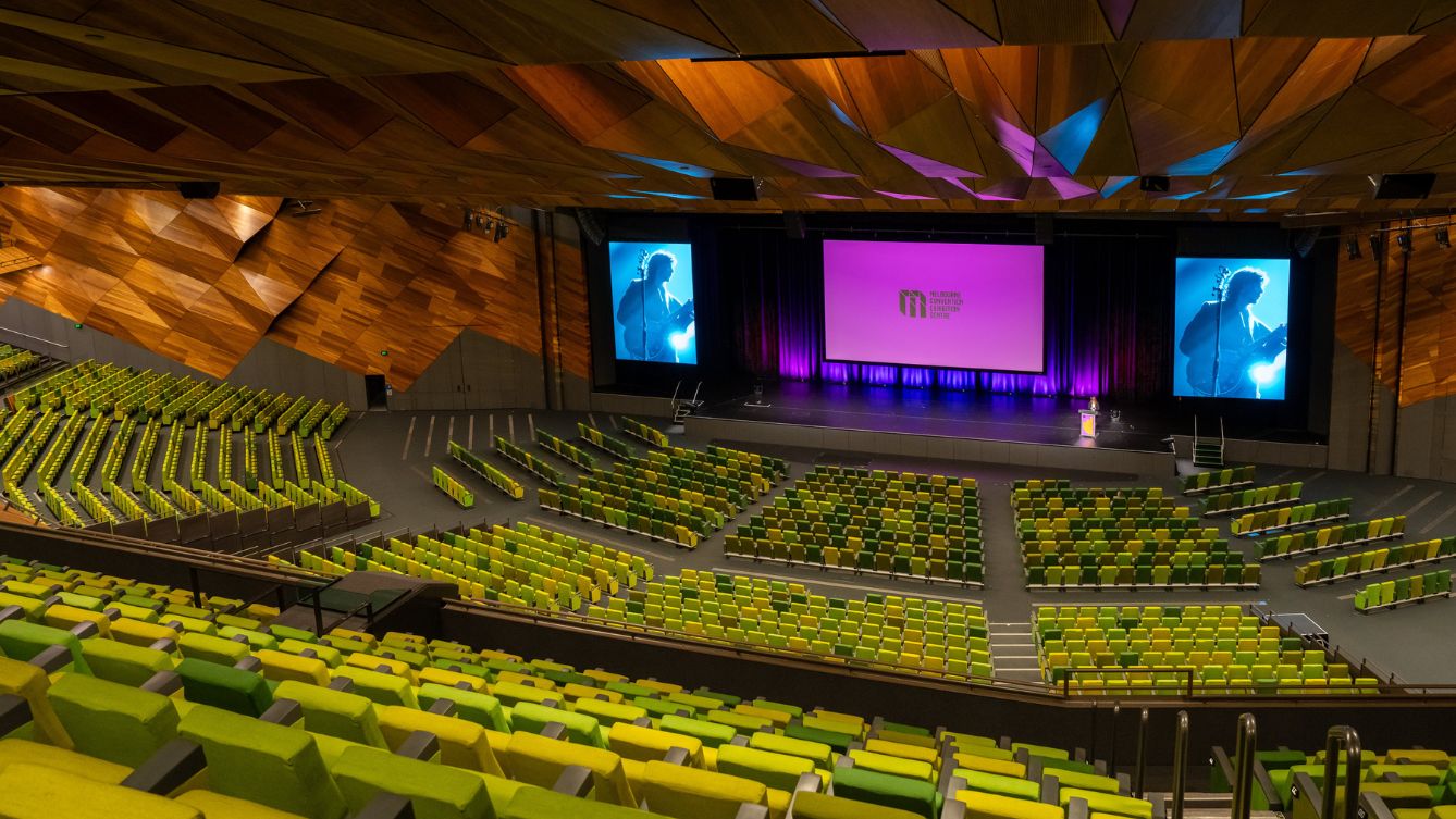 Empty view of Plenary theatre at MCEC. A large screen with a pink graphic and the MCEC logo is displayed on the screen over the stage. 