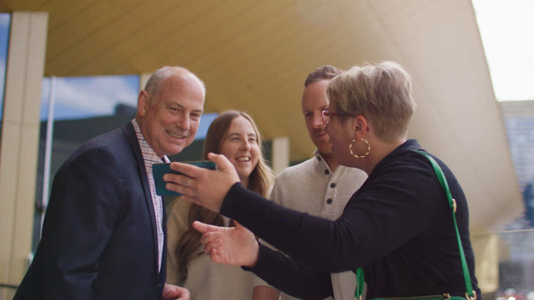 A woman holding her phone and showing the screen to three people. One of the individuals is David Howie.