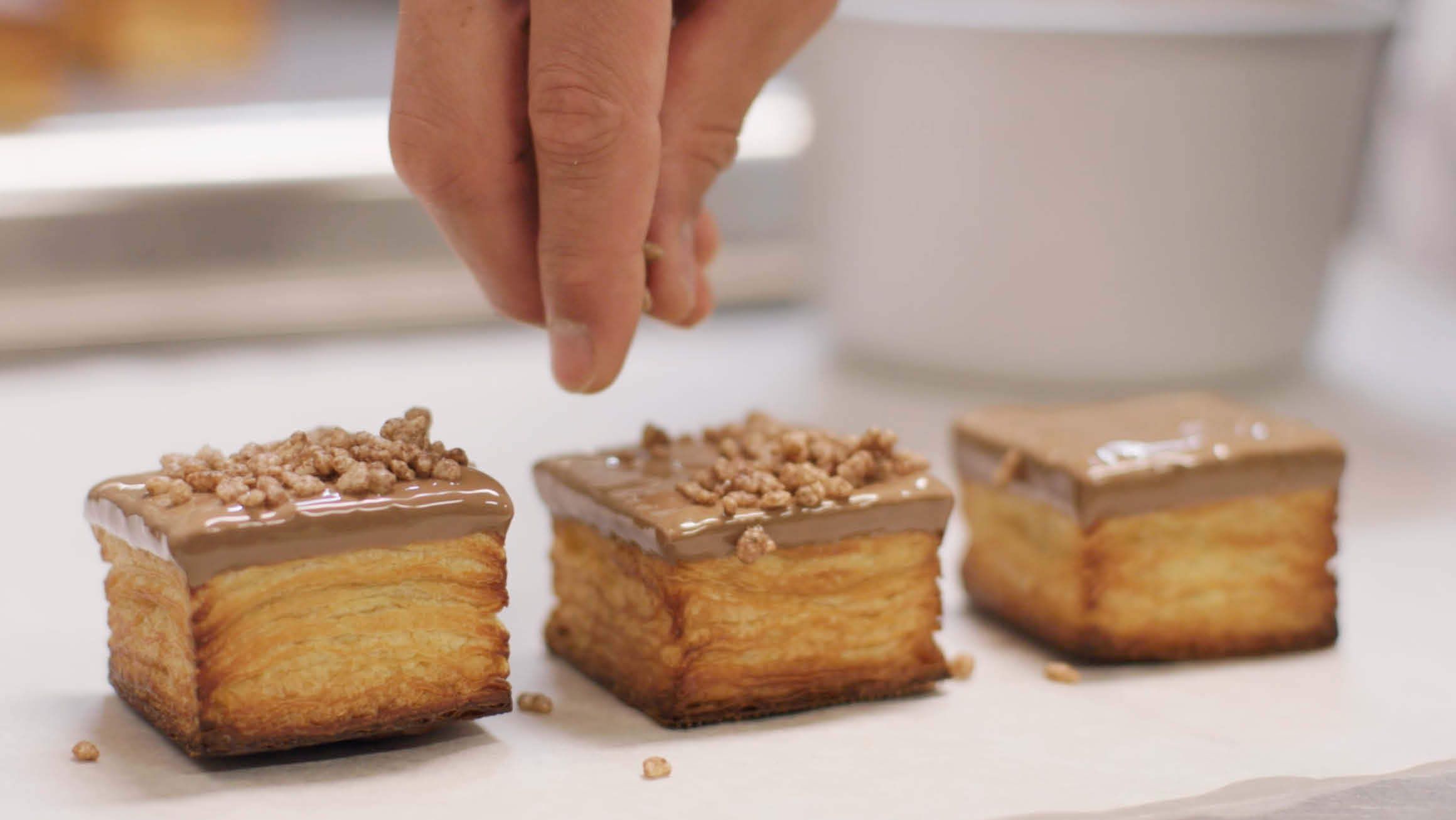 Three cakes in a row with the finger tips of a hand, off camera, sprinkling nuts on top of the chocolate icing. 