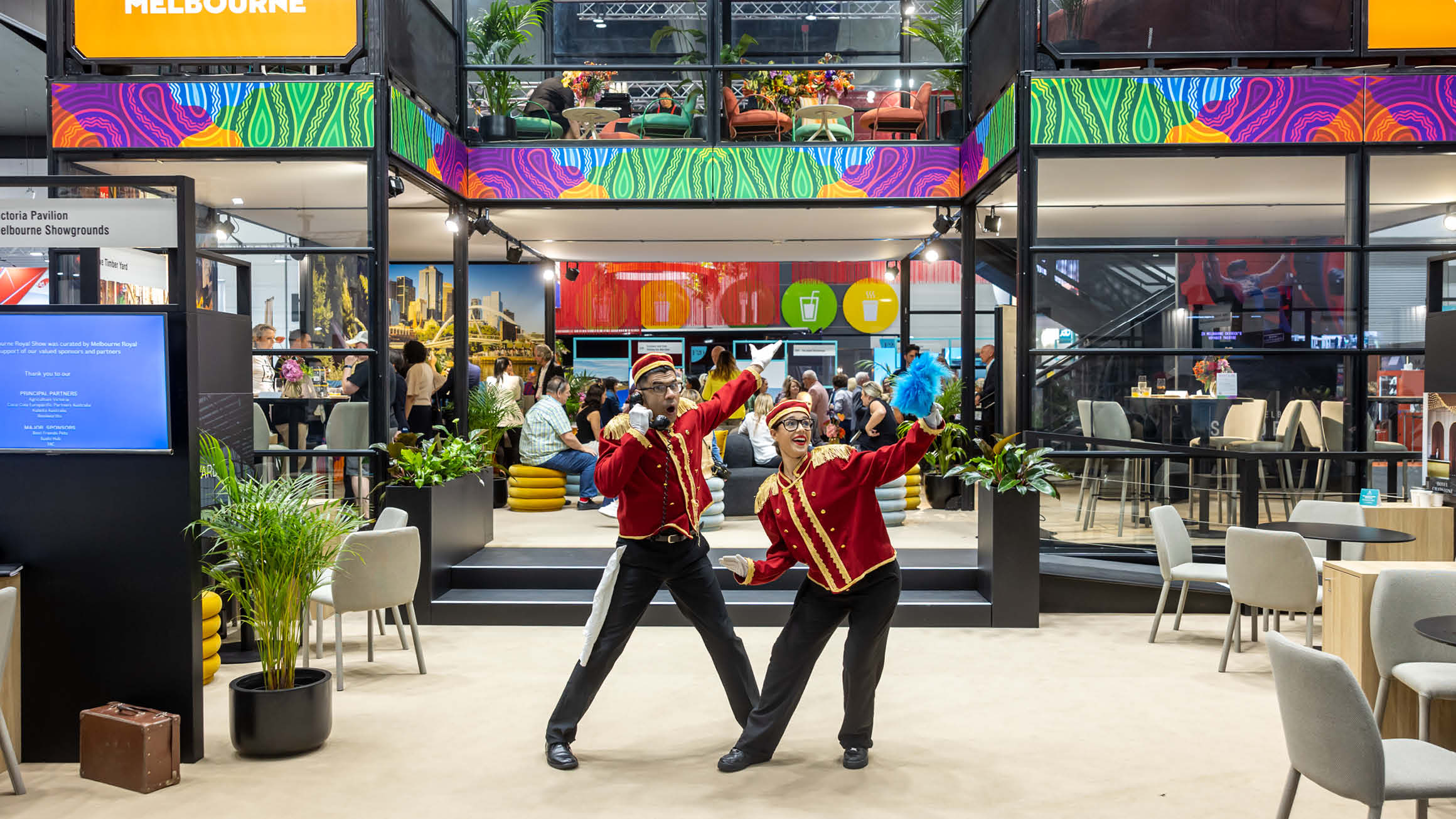 Two people dressed in bellboy costumes stand in front of a double story exhibition stand with lots of chairs and tables on both levels. 