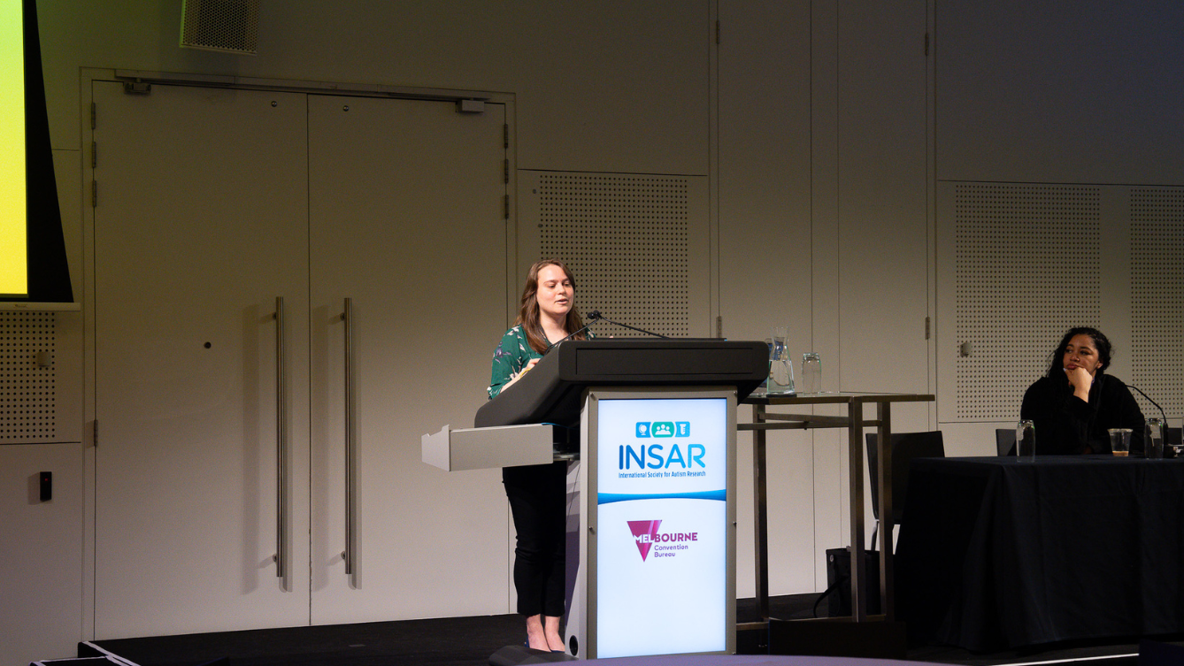 A lady stands on a stage speaking in front of a podium with the logos INSAR and Melbourne Convention Bureau displayed on the front. 