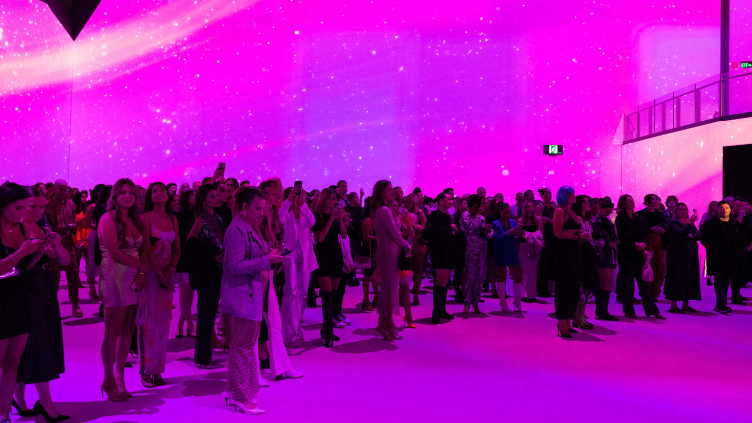 Crowd of people standing together facing a stage off camera under pink lighting. 
