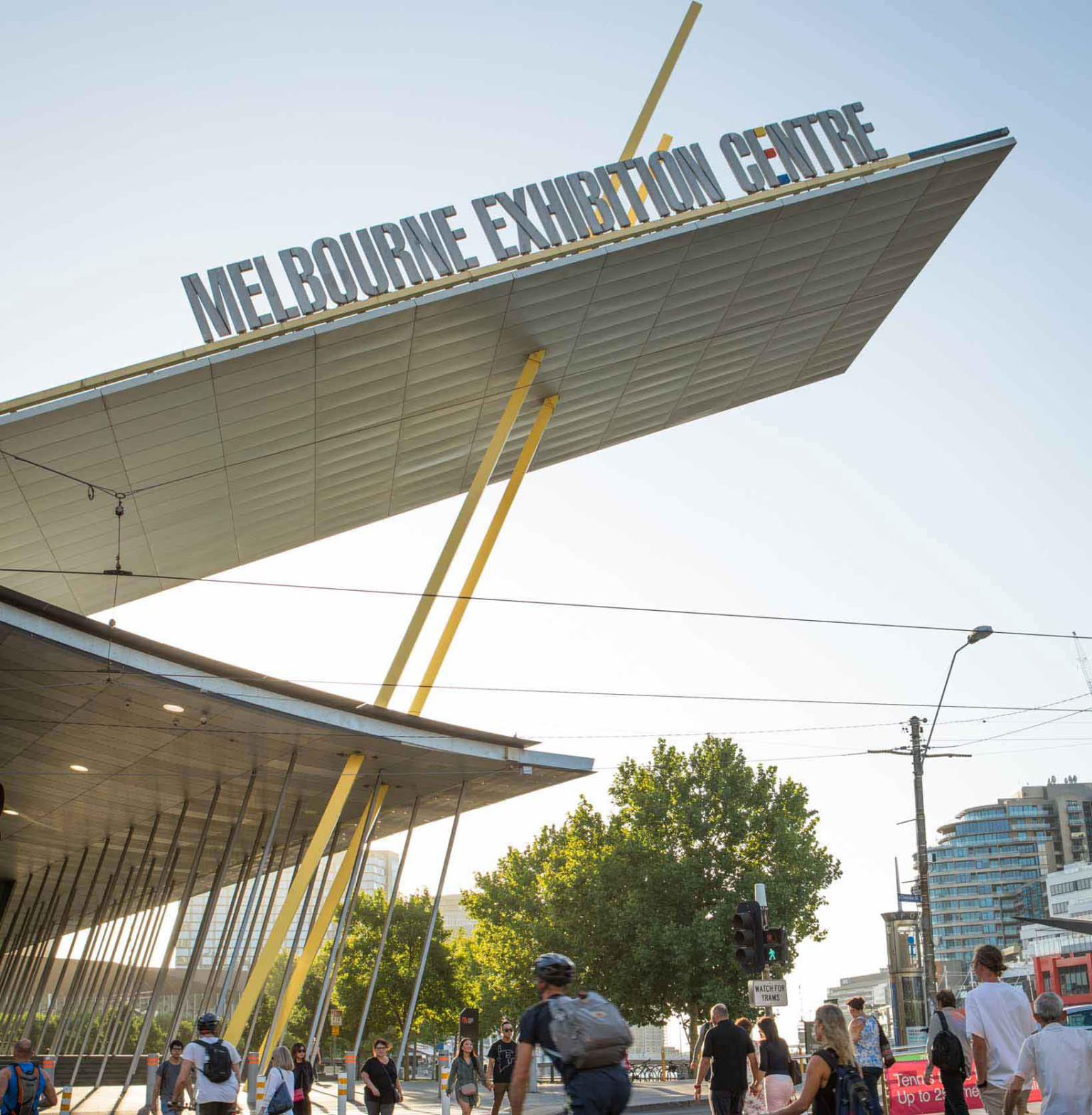 Outside view of the Melbourne Exhibition Centre with blue skies behind. People and bicyclists in the foreground are crossing the road. 