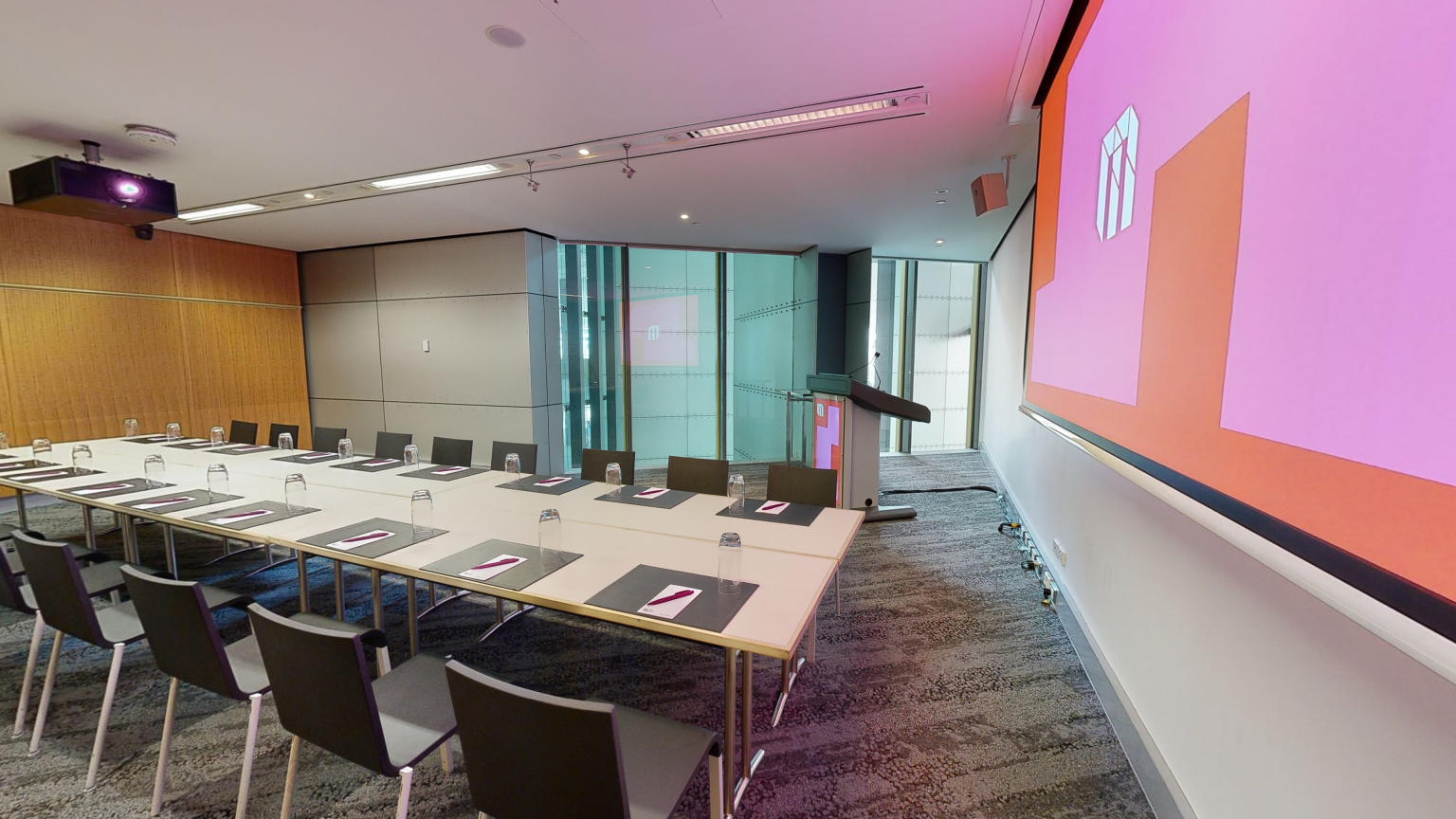 A conference or meeting room featuring a spacious boardroom layout equipped with a sizable screen and comfortable chairs for attendees. A lectern also sits in the corner to the left of the screen next to a large floor to ceiling window. 
