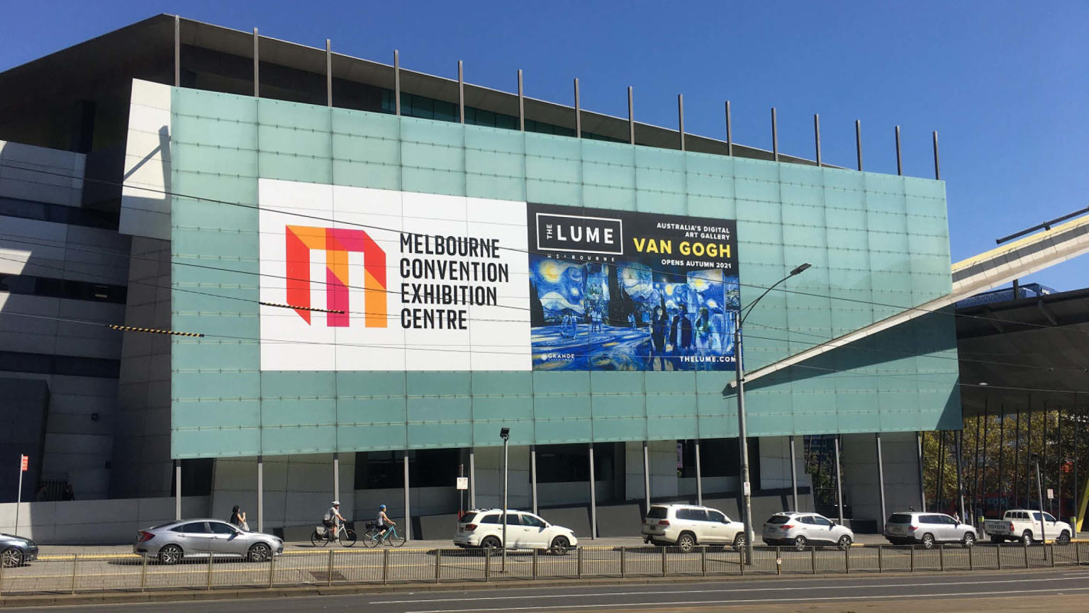 A spacious building with a prominent glass pane in front adorned with display stickers showcasing the names "Melbourne Convention Exhibition Centre" and "THE LUME Melbourne Van Gogh." Several cars are parked in front of the building, while the sky above shines in a clear, vibrant blue.