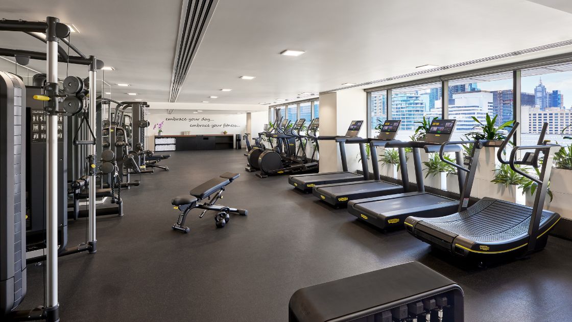 Inside the gym at Pan Pacific Melbourne. Rows of workout machines and treadmills facing a large floor to ceiling window. 