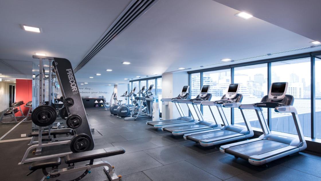 Inside the gym at Pan Pacific Melbourne. Rows of workout machines and treadmills facing a large floor to ceiling window. 