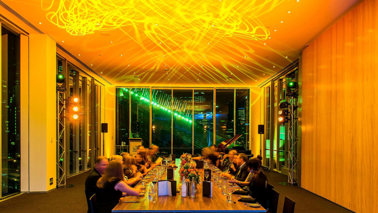 Long table with people sitting along either side in front of floor to ceiling windows. People sit at the table eating dinner with yellow lighting filling the room. 