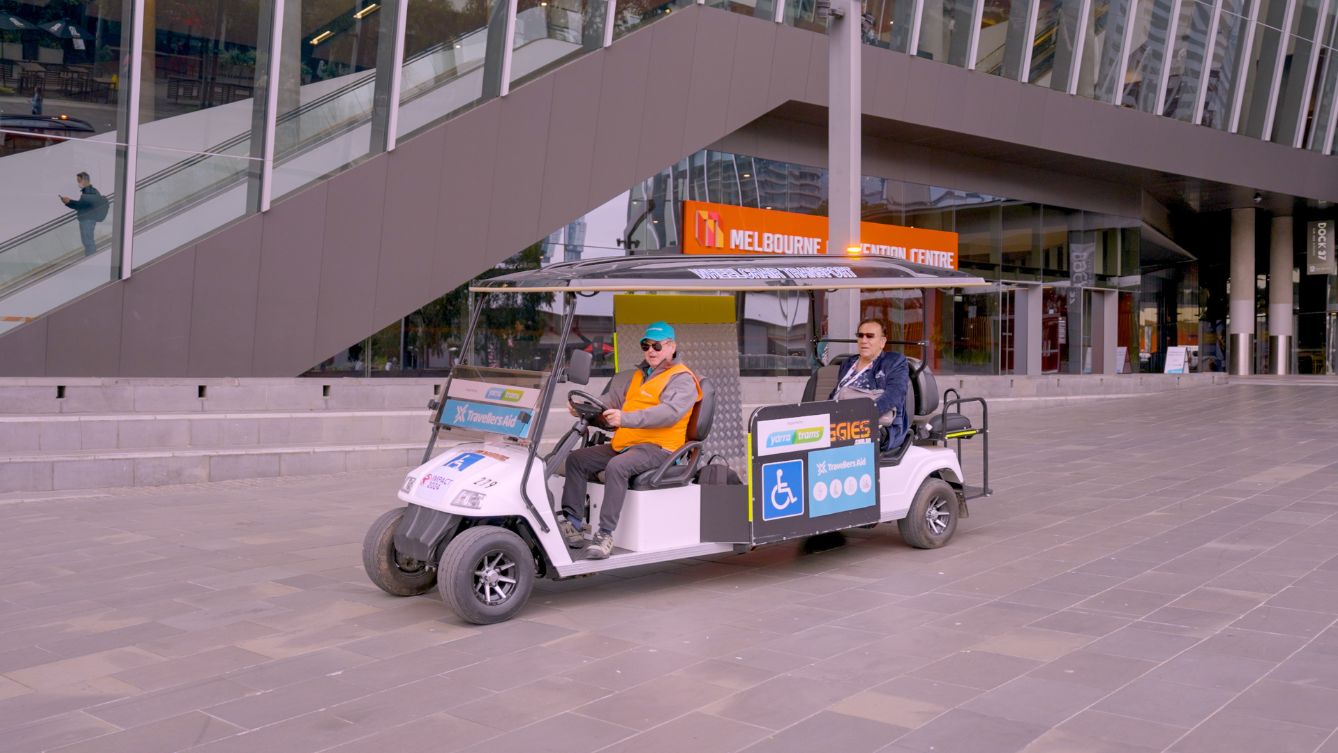 A driver in a high-vis orange vest driving a travellers aid cart with a passenger in the back outside MCEC. 