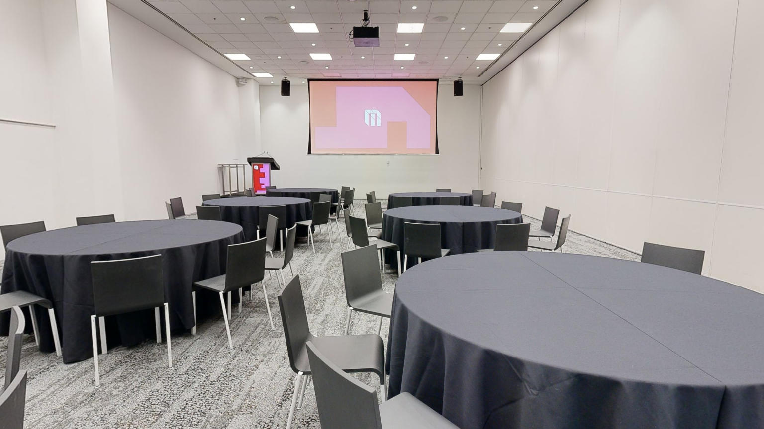 A professional conference or meeting room with neatly arranged round tables and chairs facing a large projector screen at the front of the room. A white MCEC logo is displayed on the screen over a pink and red shaped background. A lectern and stage sit to the side of the screen facing out towards the tables. 