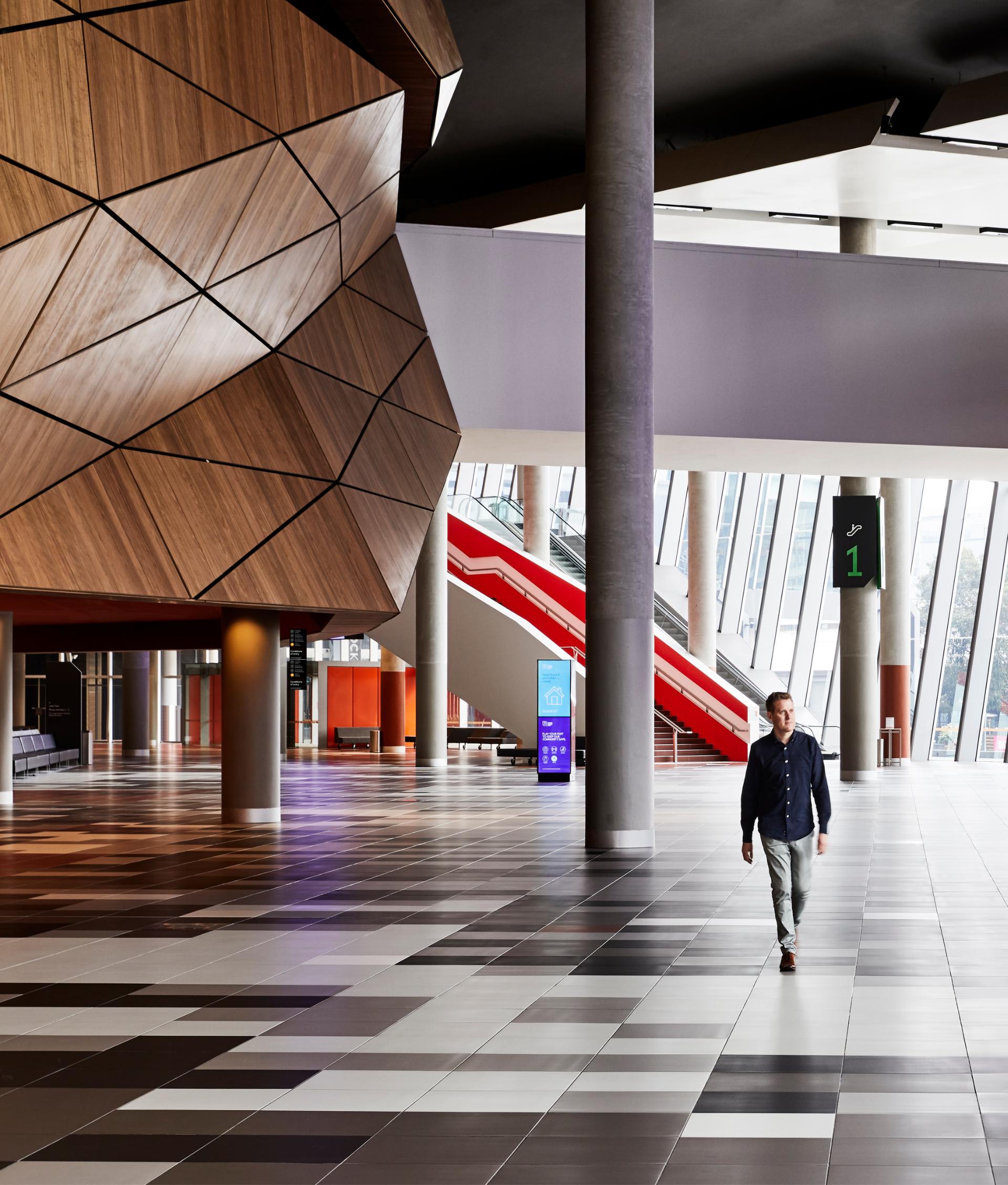 Inside view of the Convention Centre. One man walks across the black and white tile floor. Large wood panelling in an abstract shape is to the left and large windows with an escalator in front is to the right. 