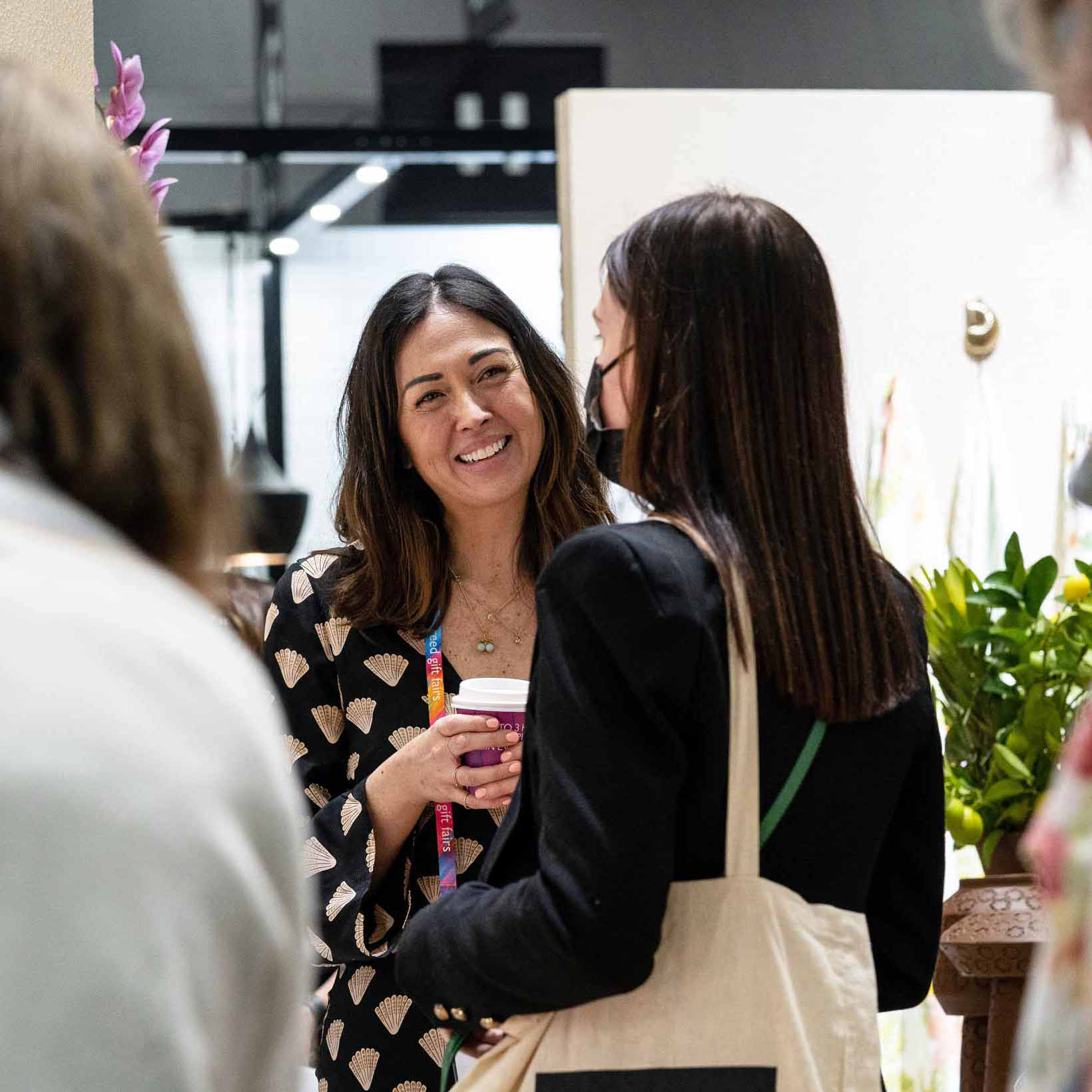 A woman holding a coffee cup smiles at another woman with her back towards the camera. The appear to be in conversation.