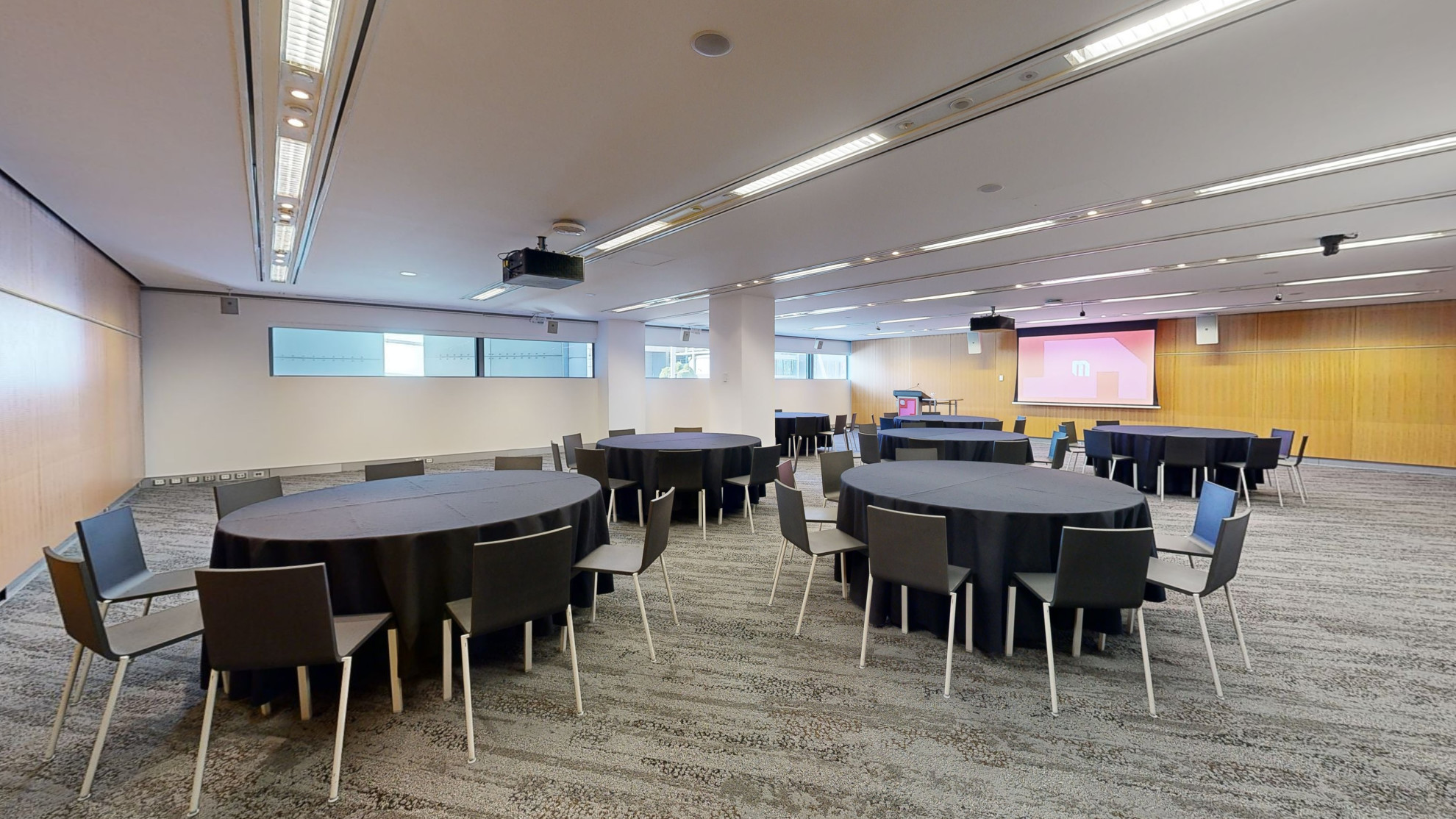 A conference or meeting room with arranged round tables and chairs for meetings or discussions. The tables focus around facing a large projector screen at the front of the room with a lectern to the left side. A strip of windows that sit at eye level run along side the far wall. 