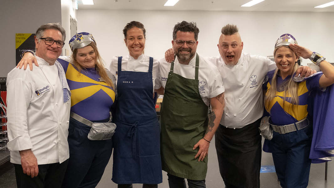 An image showing six people standing together in a line smiling at the camera. Four of them are chefs, two wearing aprons, and the other two are dressed as superheroes in purple and yellow capes with silver crowns.