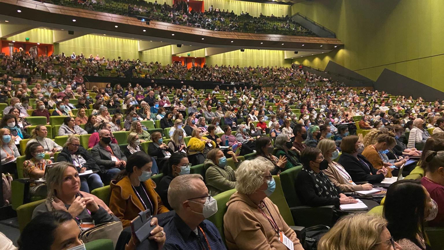 Crowd of people some with masks on sit in Plenary theatre that has green chairs and walls at MCEC. 