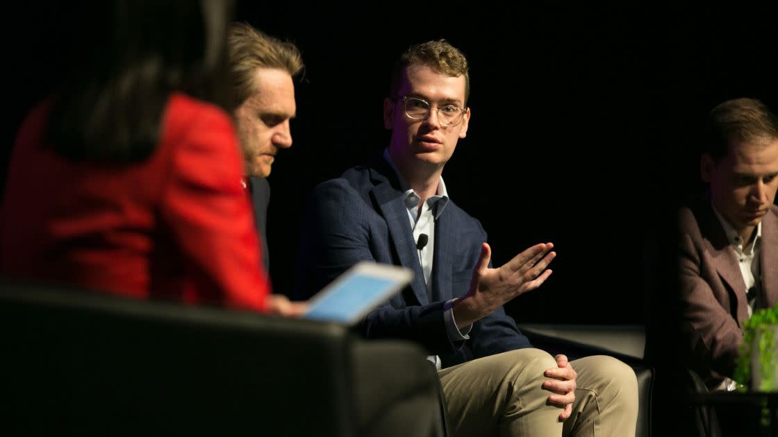 Man in jacket sitting on a panel talking to three other people on stage with him. Presenting to a crowd off camera. 
