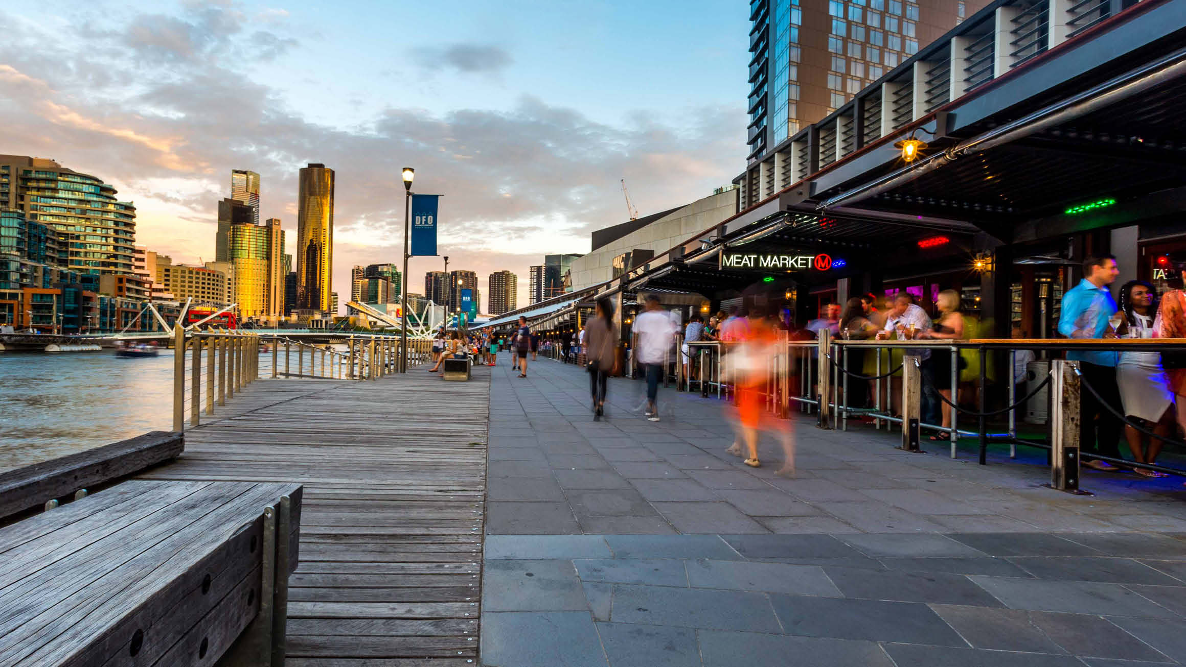 A picturesque scene capturing the South Wharf precinct in Melbourne, with the sun setting in the background. The Yarra/Birrarung River flows on the left, while people can be seen strolling along the precinct. On the right, individuals line up eagerly outside restaurants, adding to the vibrant atmosphere.