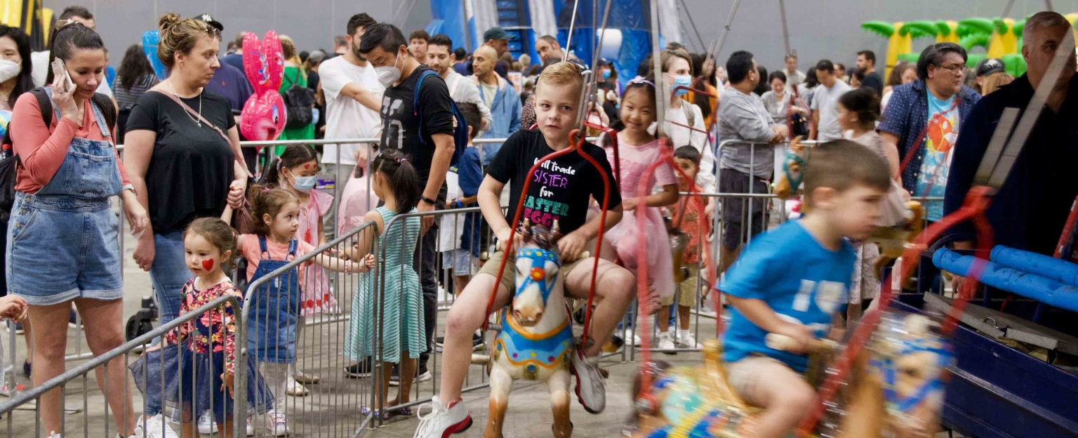 A carousel ride at the Good Friday Appeal Kid's Day Out held at Melbourne Convention and Exhibition Centre. Children are riding the horses, while other children and adults wait in line. The carousel is located in a large exhibition bay, with people walking around in the background.
