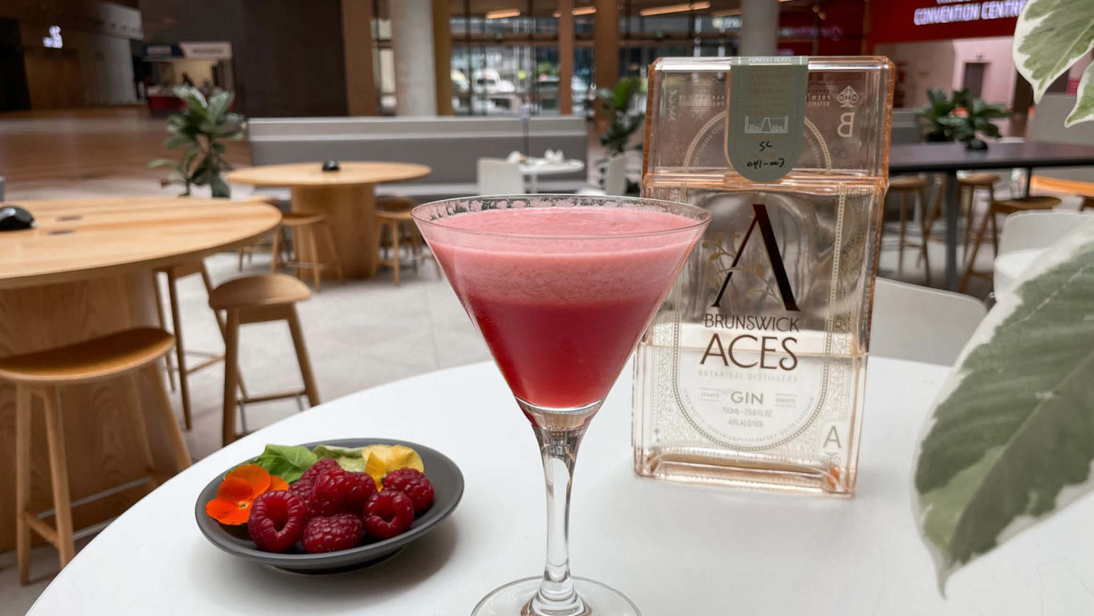 A vibrant pink cocktail fills a cocktail glass, topped with frothy foam, placed on a white round table. Adjacent to it, a small grey bowl holds raspberries, while an orange-tinted rectangular glass bottle stands nearby. In the background, round wooden tables with matching chairs create a cosy cafe atmosphere, while green leaves in the foreground add a touch of natural beauty.