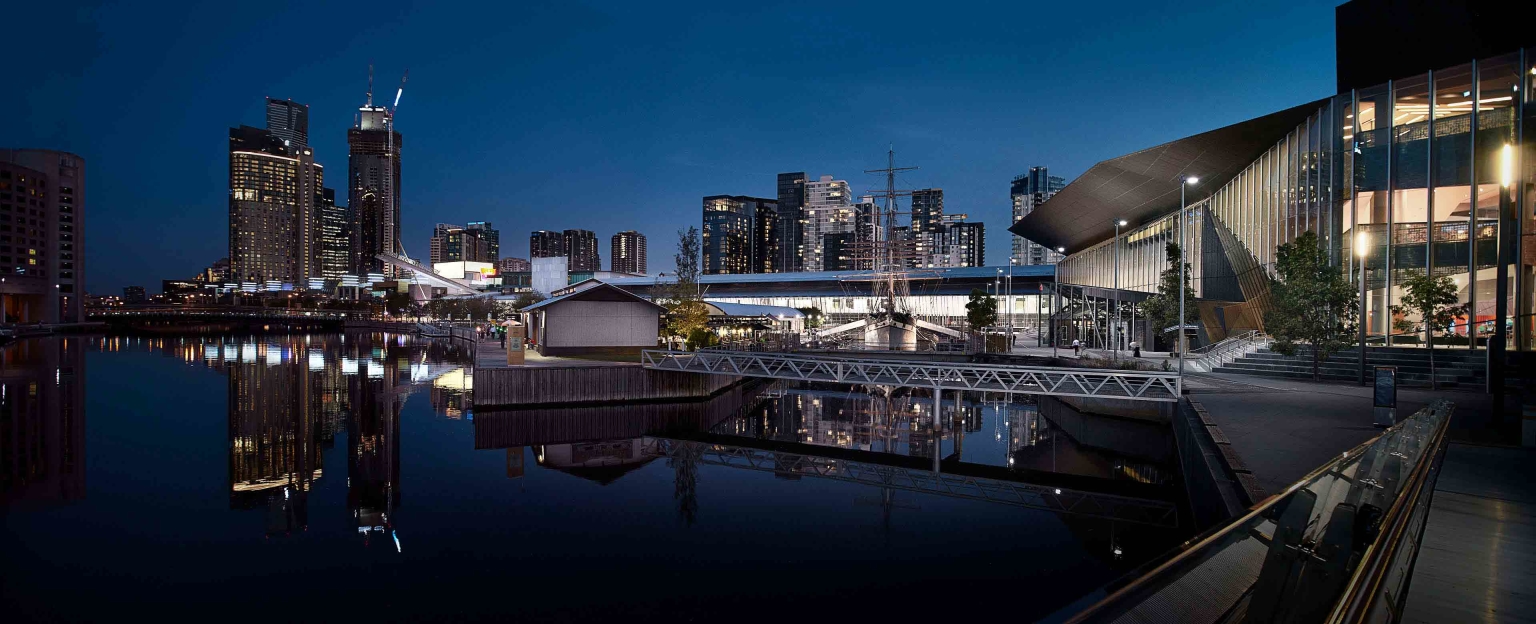 A night view of the South Wharf Promenade in Melbourne, Australia. The Melbourne Convention and Exhibition Centre and several skyscrapers are visible in the background. The Yarra/Birrarung River flows through the foreground, reflecting the lights of the city.