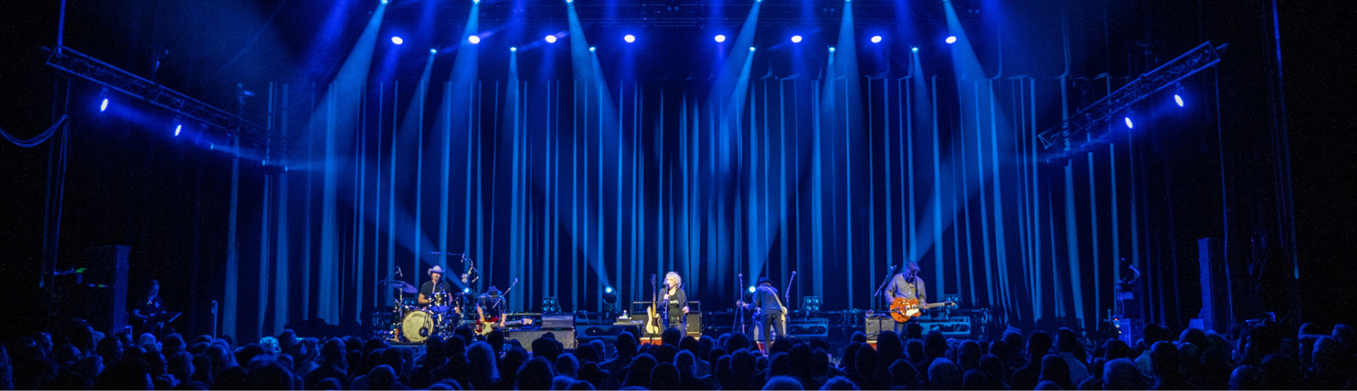 A band plays on stage with blue spotlights on them. A crowd stands under the darkness in front of them. 