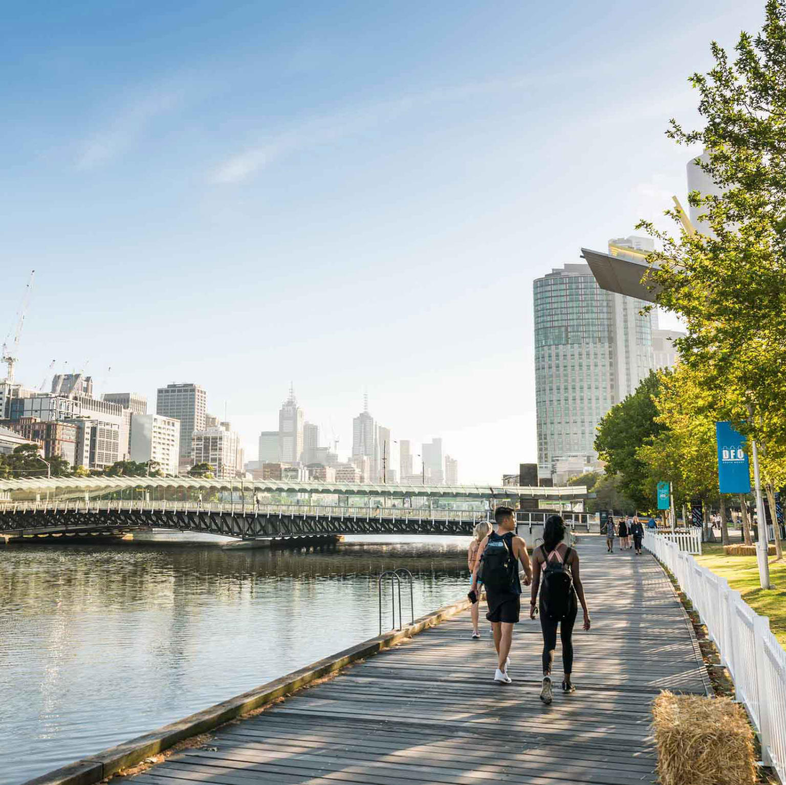 Experience Melbourne: Cityscape view, two people stroll along Yarra River's wooden walkway under a clear blue sky.
