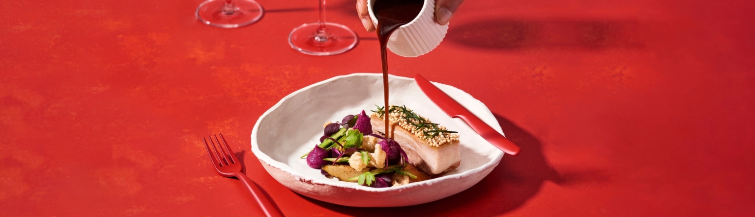 High country pork belly with red cabbage purée, spiced apple and puffed quinoa in a white bowl against a bright red background. A hand is pouring a liquid over the plate. A red knife and fork lay next to and on the bowl. 
