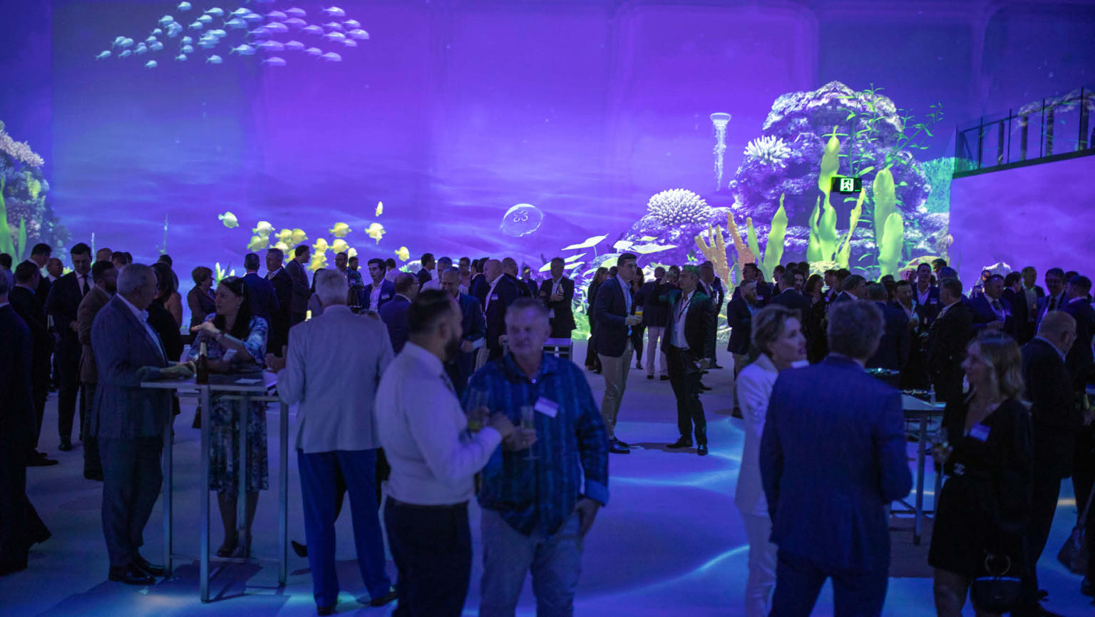 A large crowd assembled in a spacious room enveloped by expansive digital screens. The screens depict a mesmerising underwater scene with shades of purple, immersing the audience in a captivating aquatic atmosphere.
