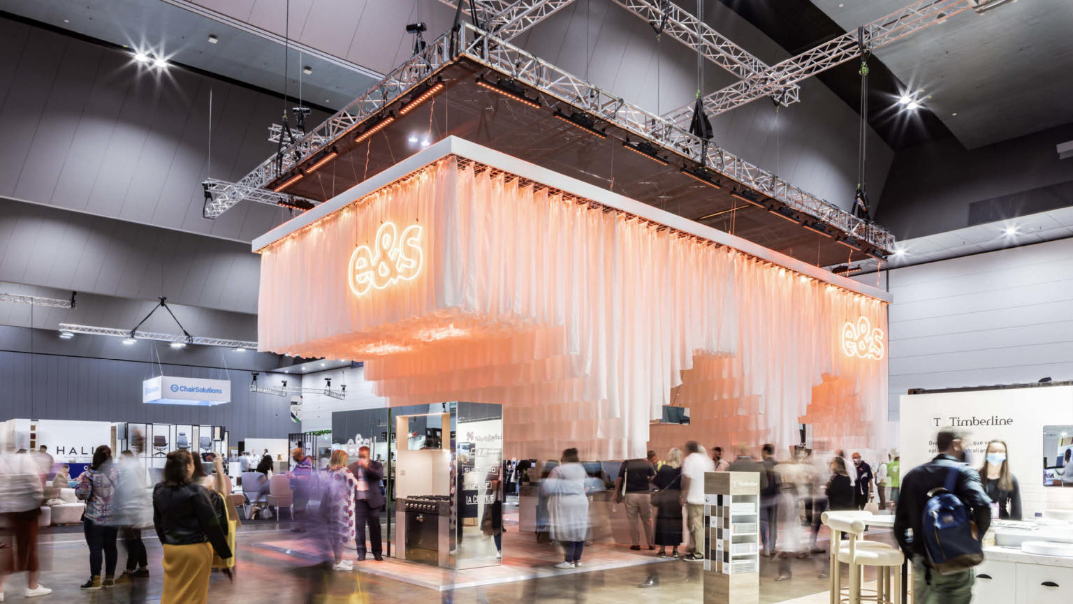 A visually stunning exhibition bay featuring a grand tiered fringe chandelier with lights and a neon logo, as people leisurely walk and explore the space.