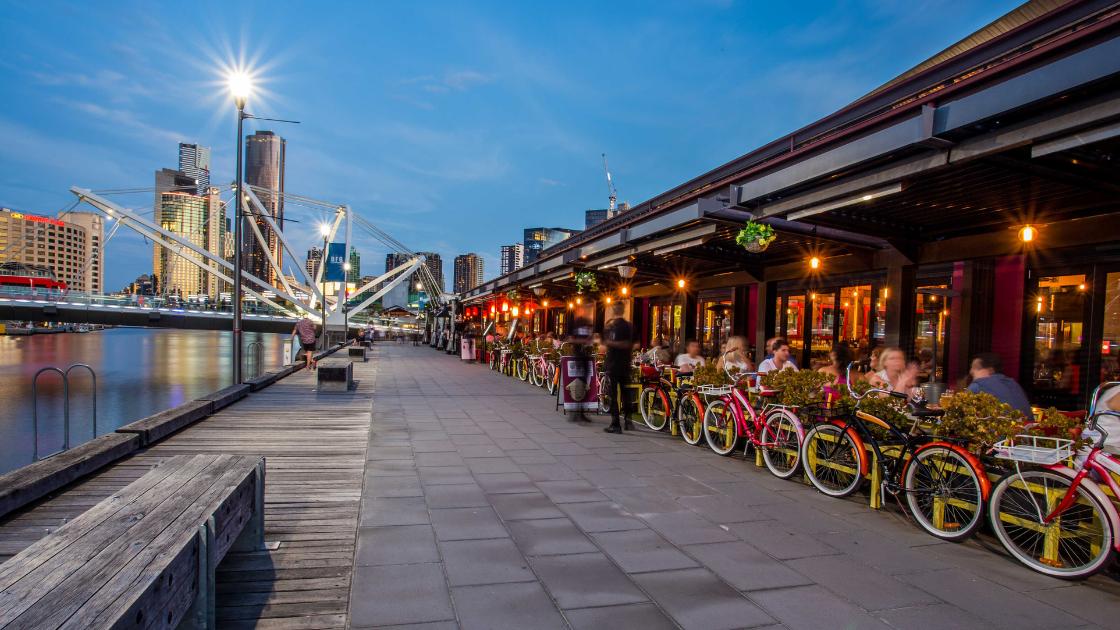 People sit at a restaurant outside along a boardwalk running along side the Yarra River. It is dusk and bright lamp shines in the corner. 