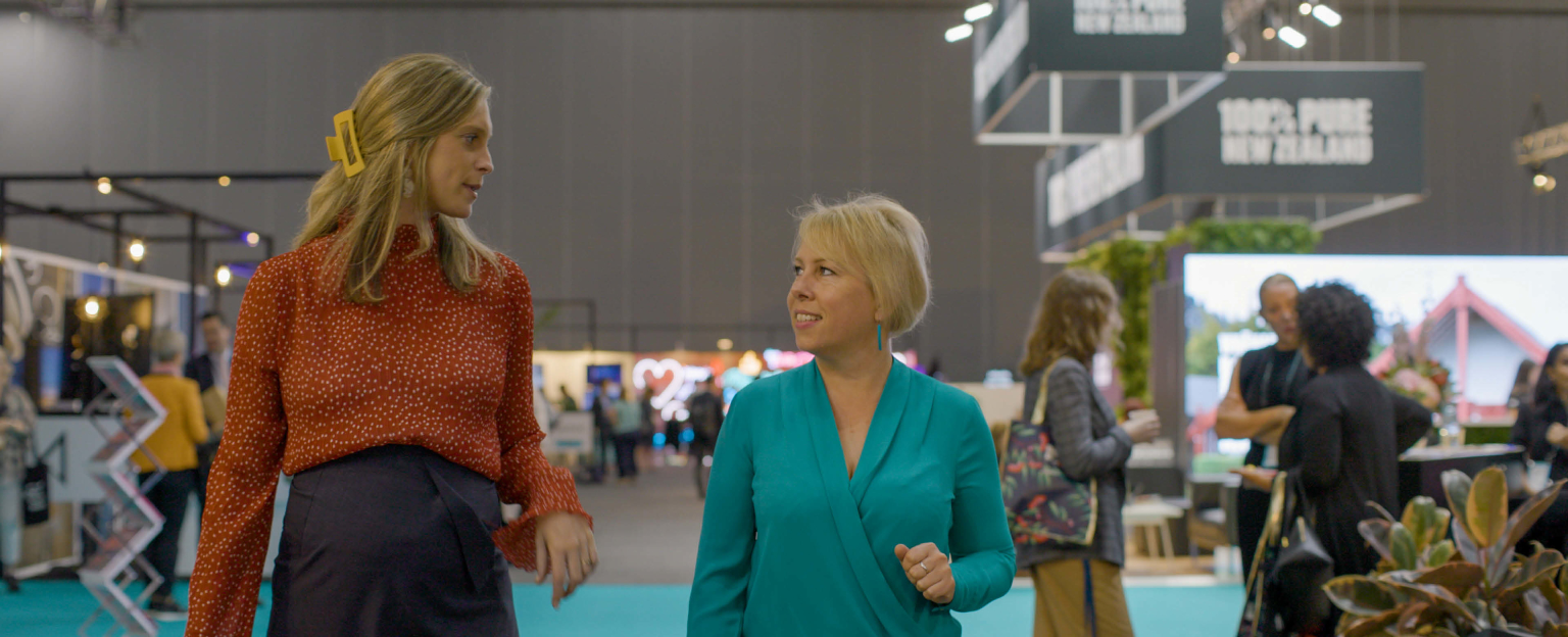 Two women, one in an orange jumper and the other in teal top are walking through an exhibition talking to one another. 