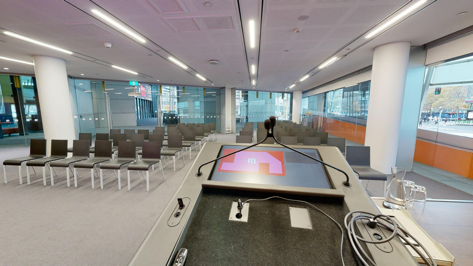 A conference or meeting room featuring rows of chairs arranged to face large tv screen for presentations and discussions. A lectern sits to the side of the tv screen and windows run along the side of the room facing out towards Crown. 