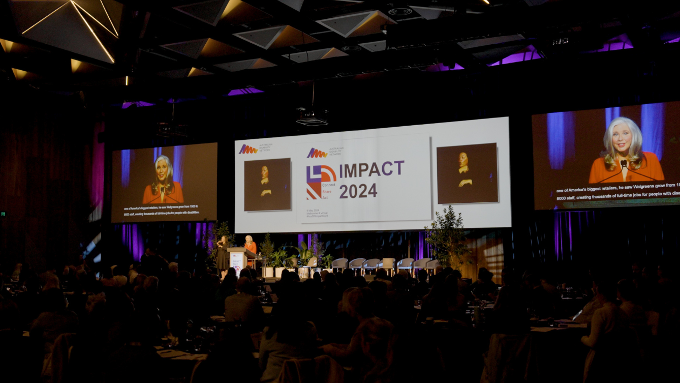 A large crowd sits in the darkness facing a stage lit up. A large screen with words IMPACT 2024 is displayed and two video screens with a woman in an orange jacket are on either side. 