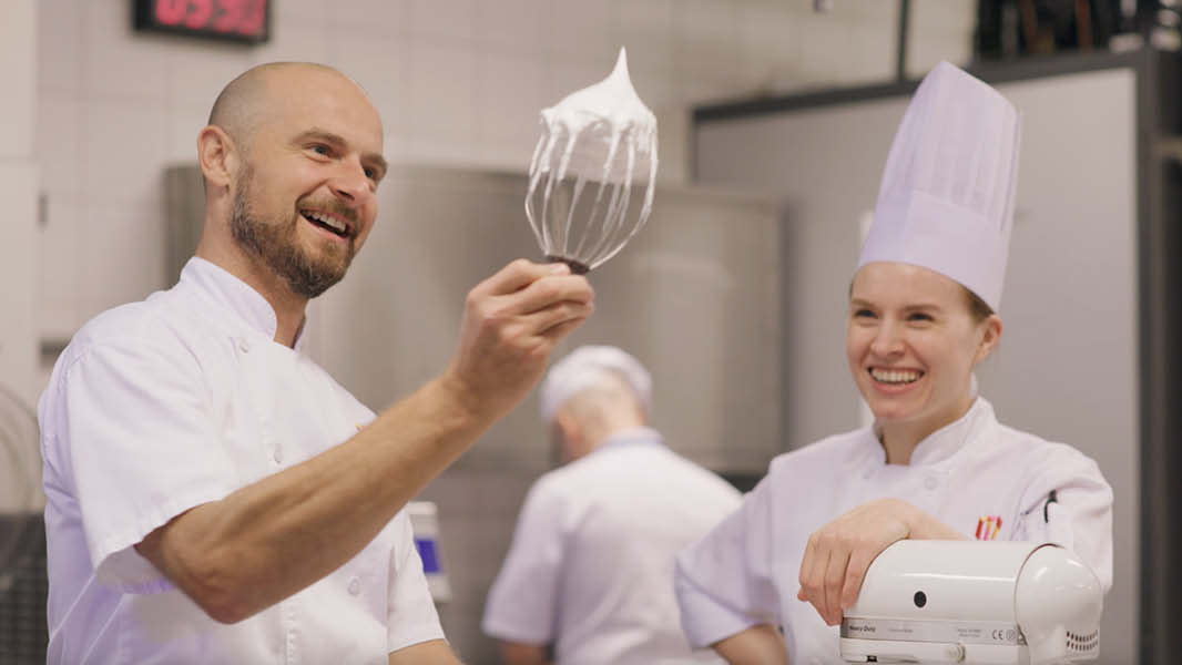 One male chef holds up a mixer head covered in cream, showing it to another female chef, who wears a MCEC (Melbourne Convention and Exhibition Centre) uniform, smiles at him. 