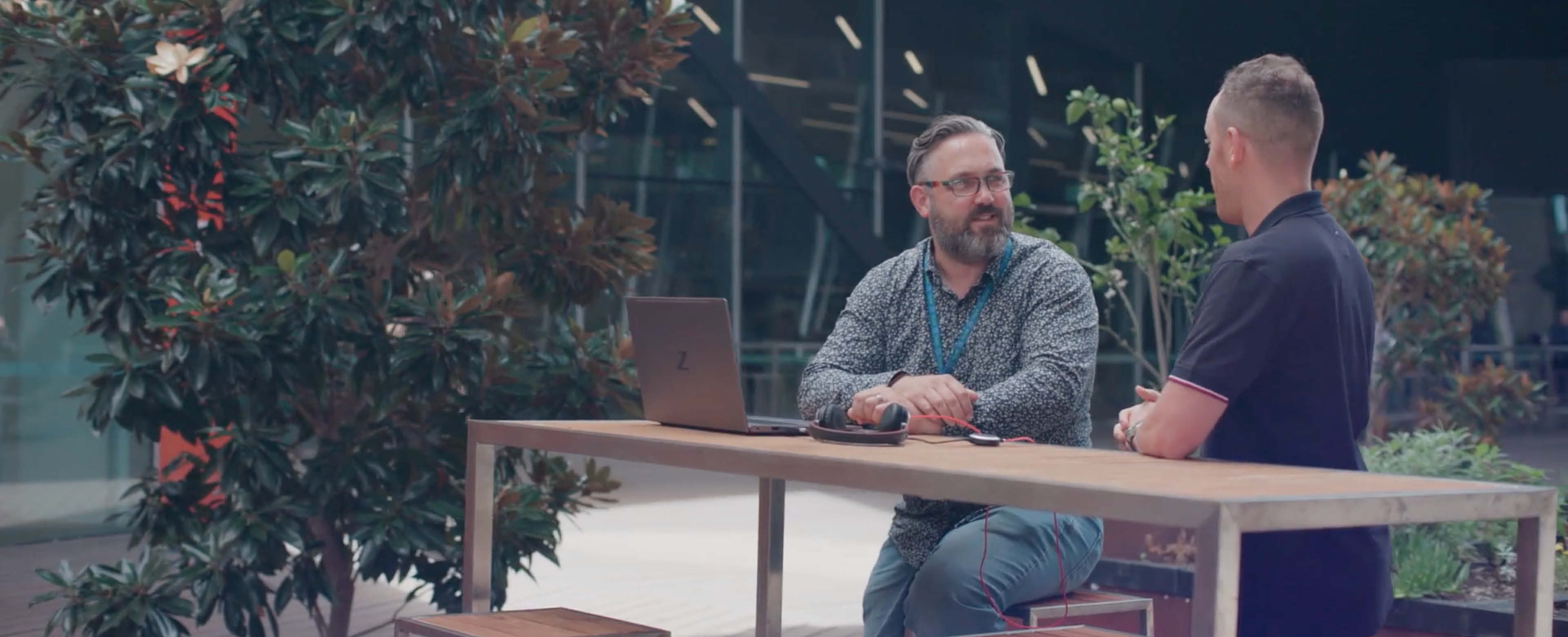 Mark Higgins, in button up shirt, sitting down a table outside talking to another man in a polo shirt. 