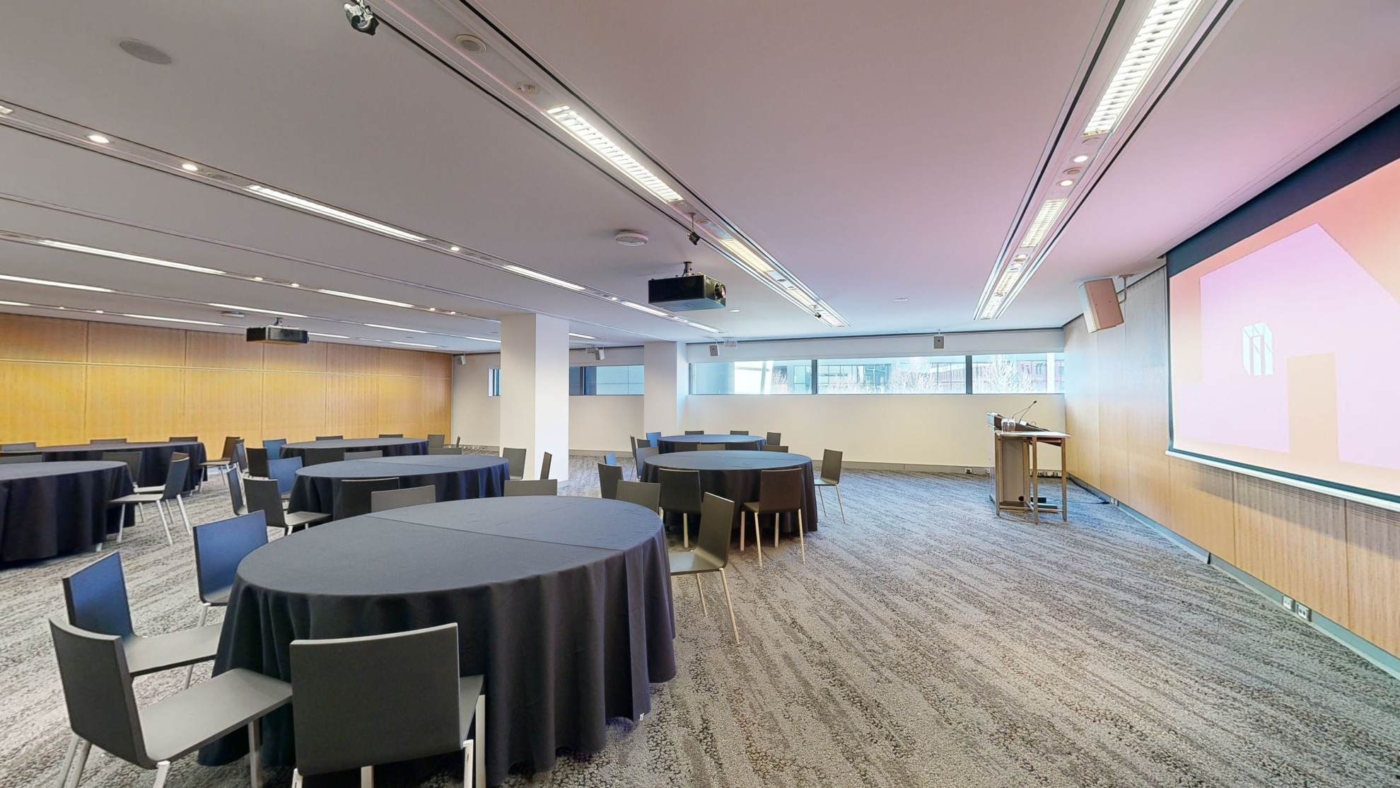 A conference or meeting room with arranged round tables and chairs for meetings or discussions. The tables focus around facing a large projector screen at the front of the room with a lectern to the left side. 