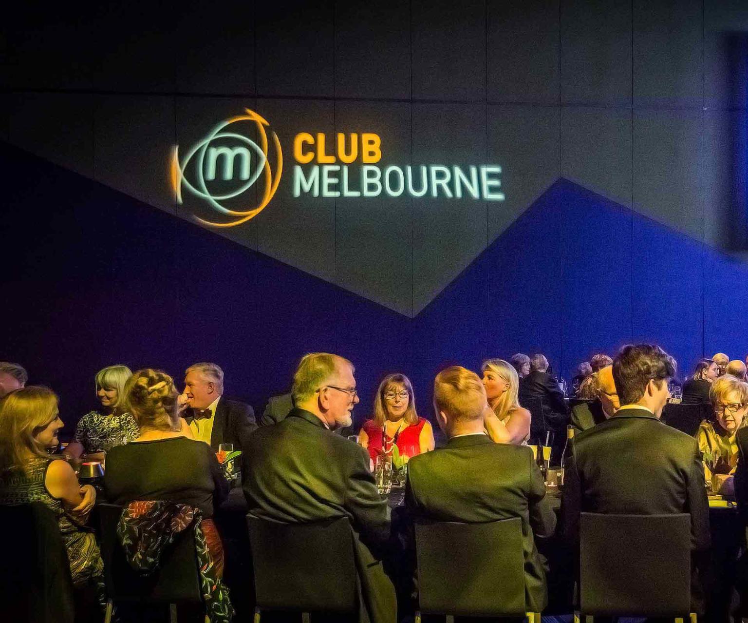Group of people sitting at a formal table talking with a Club Melbourne sign in the background on a blue and black wall. 