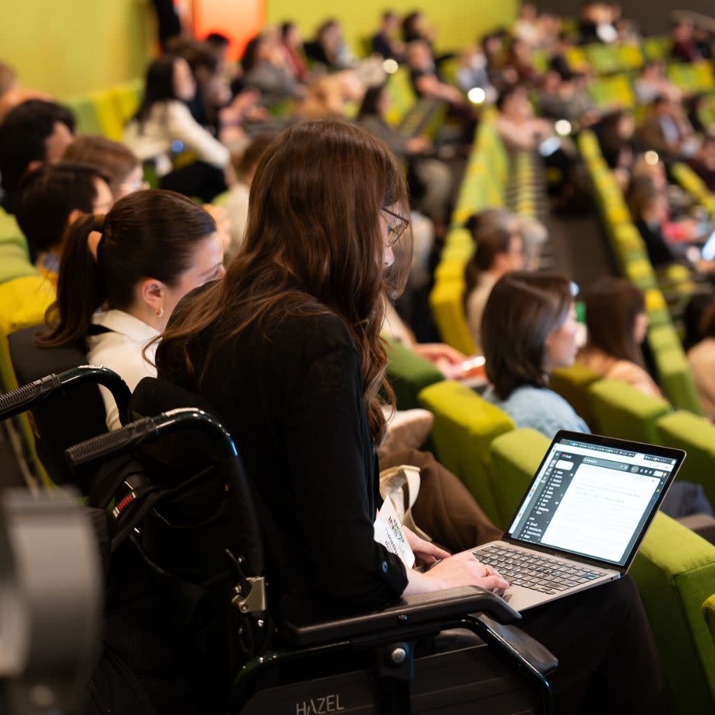 A person is sitting in a wheelchair at the end of a row of green seats in a theatre. The seats are filler with people. 