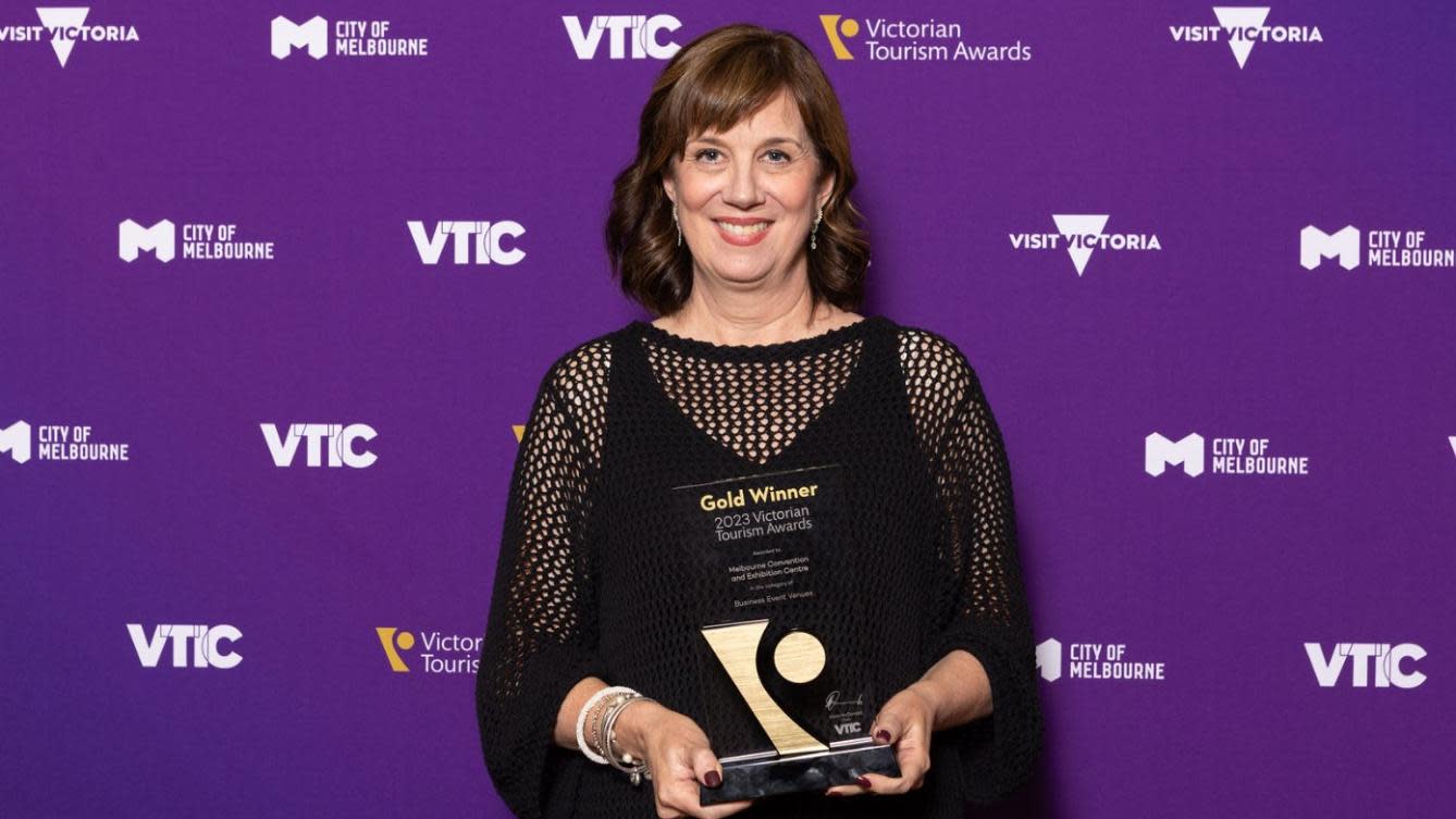 MCEC CEO, Natalie O'Brian, in a black dress standing in front of a purple background with the Visit Victoria, City of Melbourne and VTIC logos across it. She is holding a Gold Winner trophy that MCEC won for Business Event Venue of the Year.
