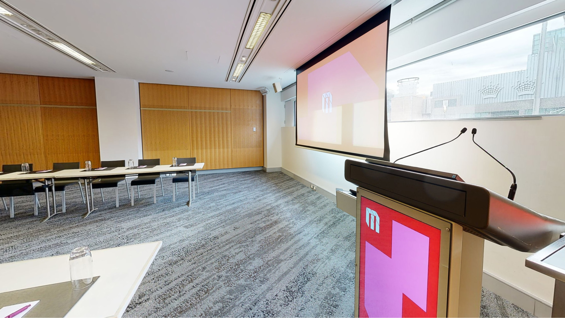 A meeting or conference room with tables and chairs arranged in a u-shaped boardroom configuration. The tables and chairs are facing a large projector screen and lectern both which have MCEC branding displayed on it. 