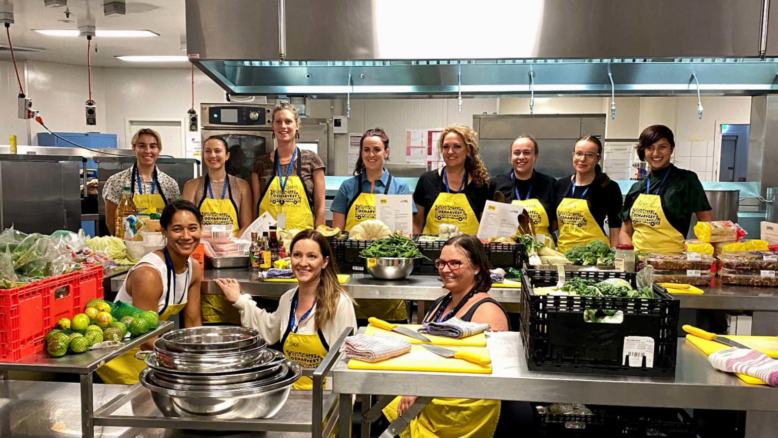 An image showing a group of 11 people standing in a commercial kitchen surrounded by ingredients, all wearing yellow OzHarvest aprons. 