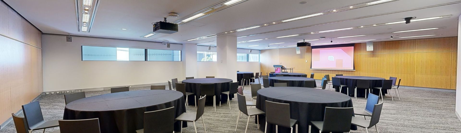 A conference or meeting room with arranged round tables and chairs for meetings or discussions. The tables focus around facing a large projector screen at the front of the room with a lectern to the left side. A strip of windows that sit at eye level run along side the far wall. 