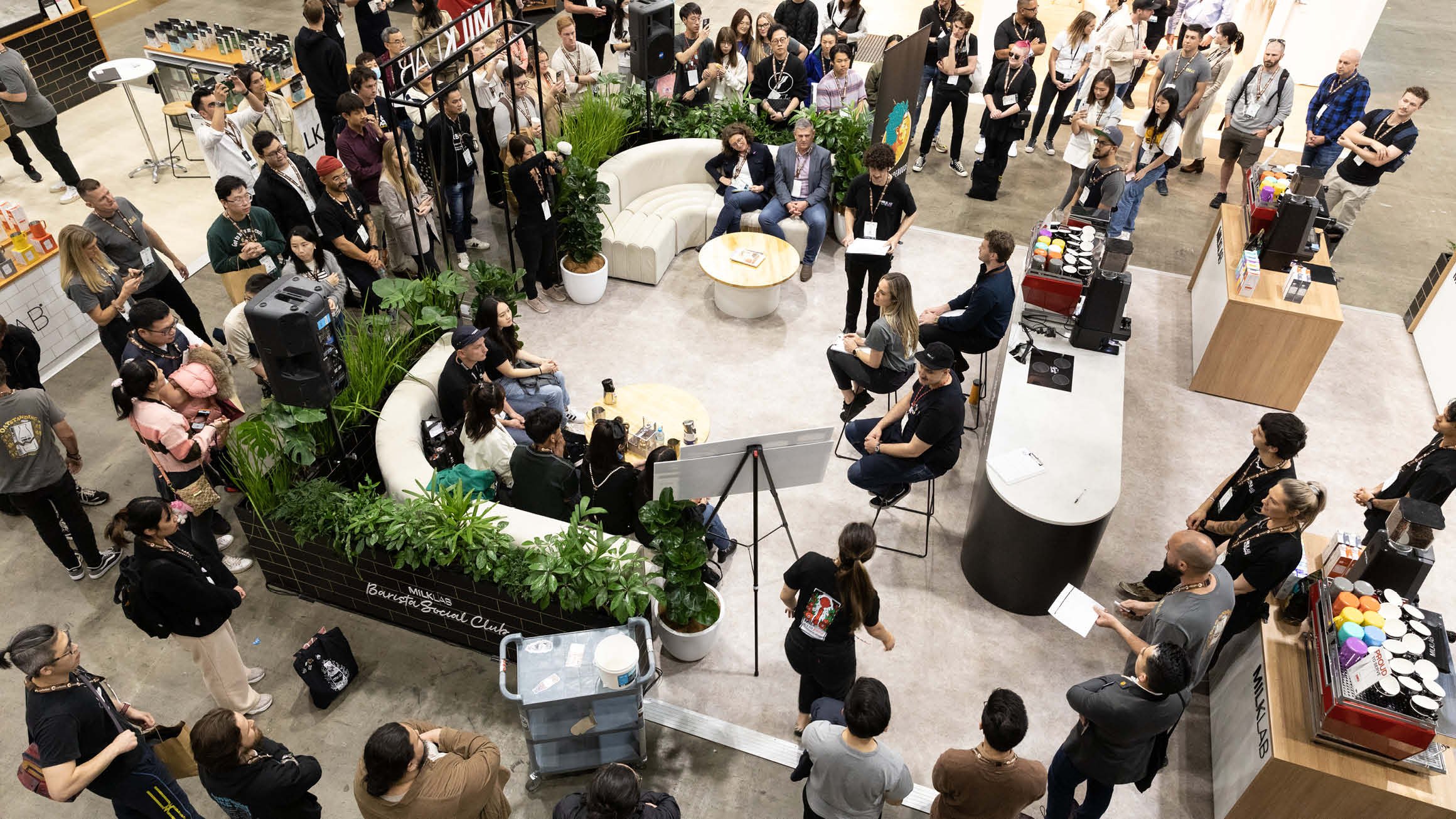 An aerial view of a spacious gathering, with individuals seated and standing around. Some relax on white couches adorned with greenery, while others stand on the outskirts, all facing a panel of three individuals.