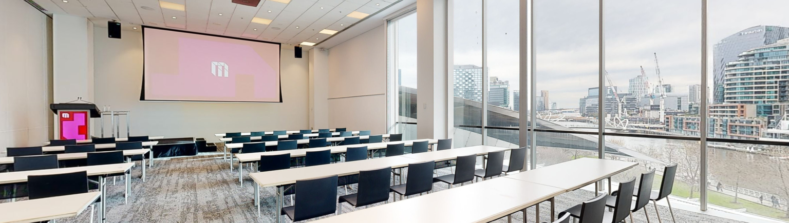 A professional conference or meeting room featuring a spacious layout, equipped with a large screen and comfortable chairs for meetings and presentations. A large floor to ceiling window fills one wall of the room, which looks out to the Melbourne city. 