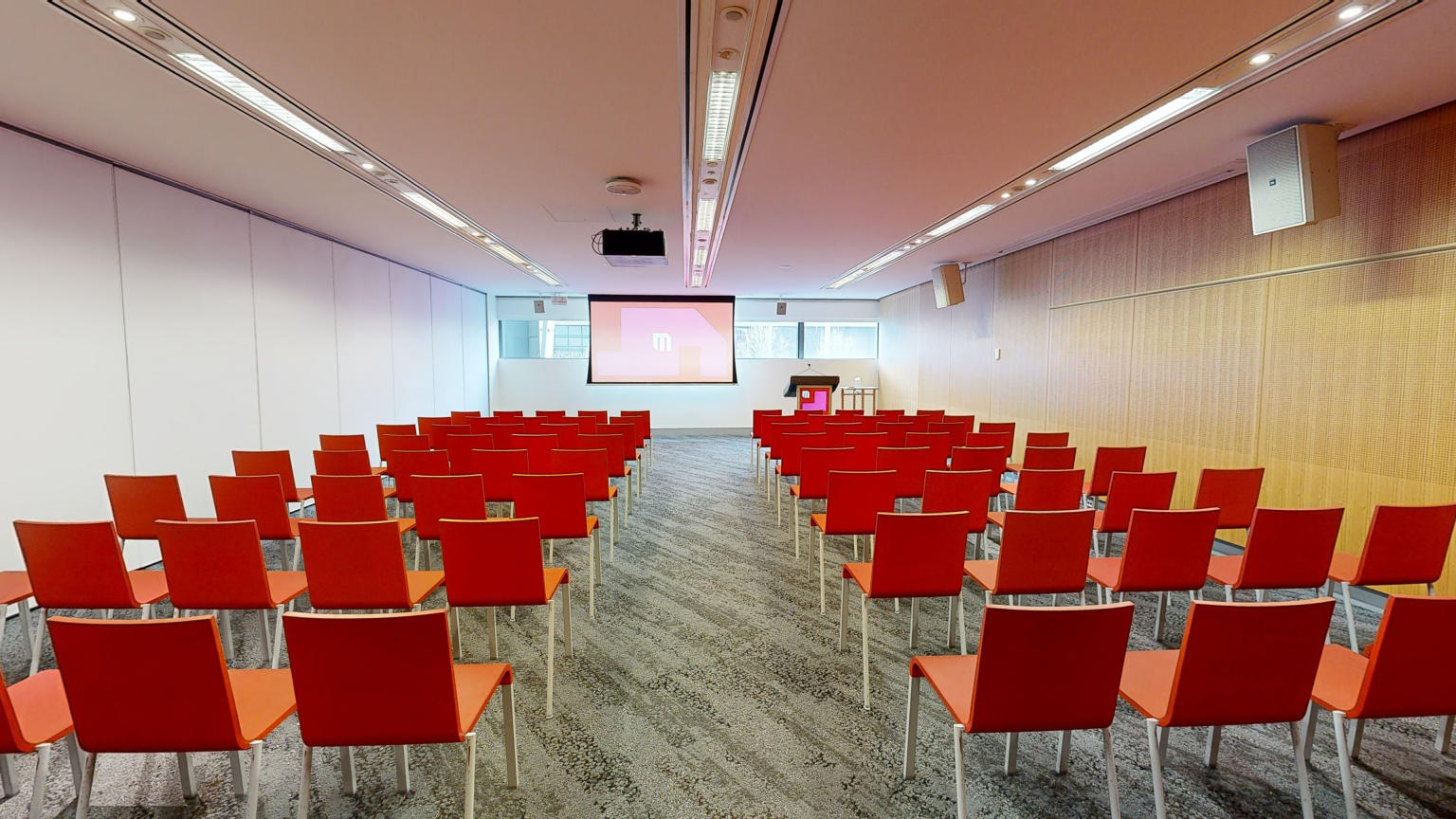 Conference or meeting room with orange chairs set up in rows facing a large projector screen with a strip of windows sitting behind at eye level. A lectern sits to the right of the screen. 