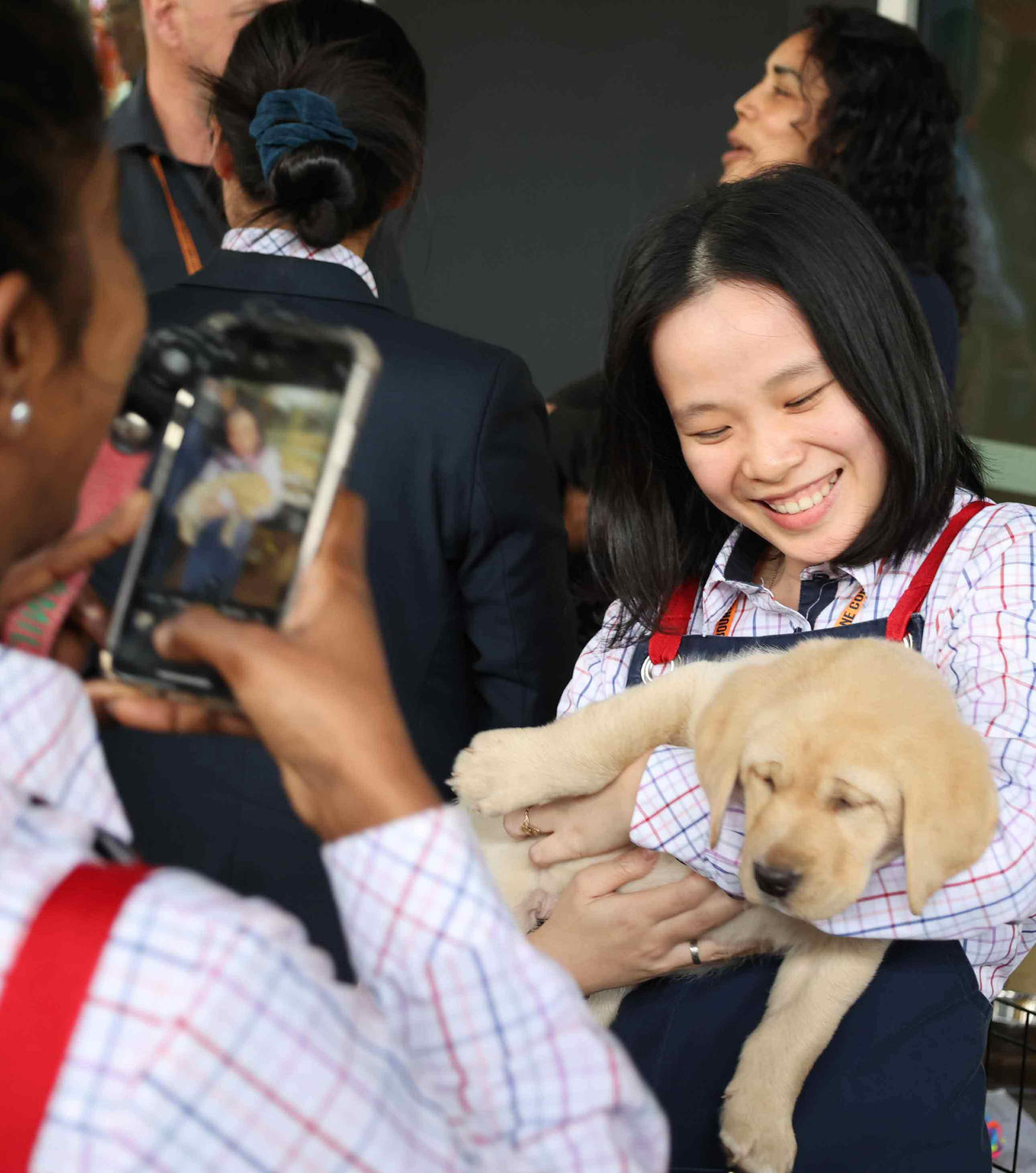 A woman holding a golden retriever puppy is smiling while looking at the puppy. Another woman in the foreground is seen taking a picture of them. 