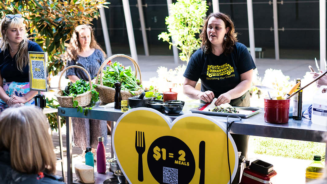 An image showing a woman demonstrating cooking a meal at a table outdoors surrounded by produce, with a group of people standing around the table and watching with curiosity. 