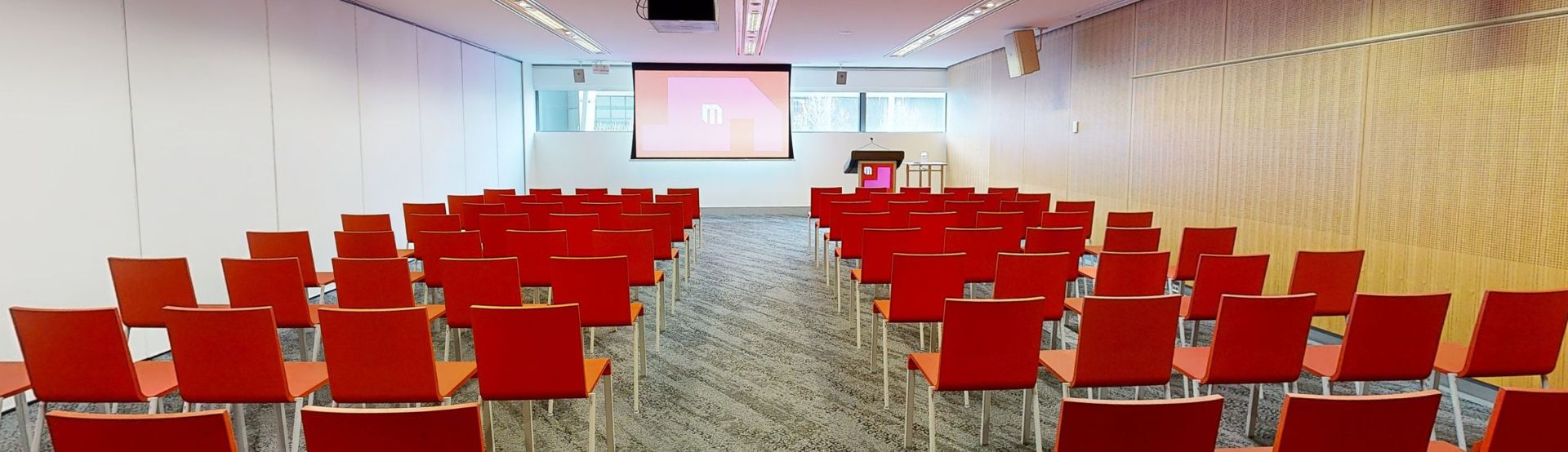 Conference or meeting room with orange chairs set up in rows facing a large projector screen with a strip of windows sitting behind at eye level. A lectern sits to the right of the screen. 