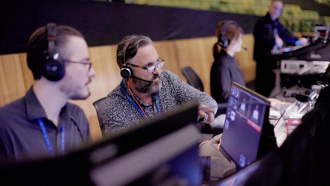 Mark Higgins, in a shirt with headphones on, talking to another man sitting next to him while pointing at a screen at a technology desk. 